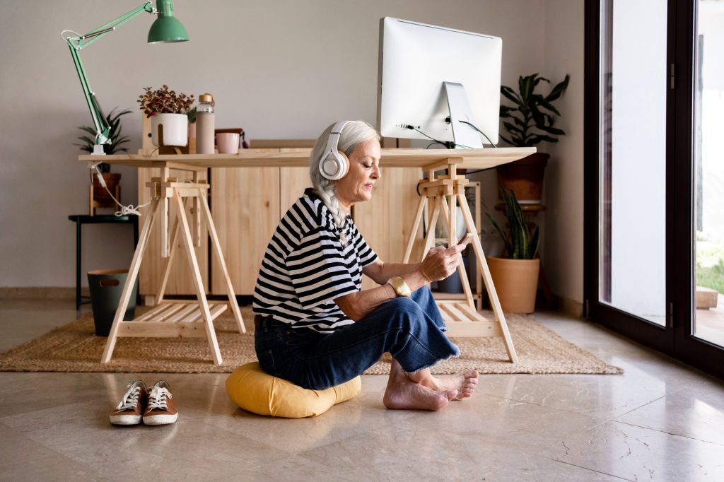 Relaxed senior woman sitting on the floor doing meditation using headphones at office